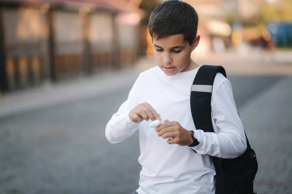 Tienerjongen pakt een draadloze koptelefoon en stopt die in de oren. Jonge jongen in wit shirt met rugzak. Jongen luister muziek — Stockfoto