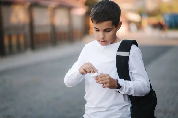 Un adolescente saca unos auriculares inalámbricos y ponlos en las orejas. Niño de camisa blanca con mochila. Boy escuchar música — Foto de Stock