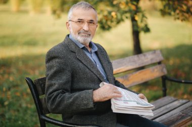Sonbahar parkında gözlüklü, sakallı, yaşlı bir adam gazete okuyor. Yakışıklı, gri saçlı adam sabahın köründe bankta oturuyor.