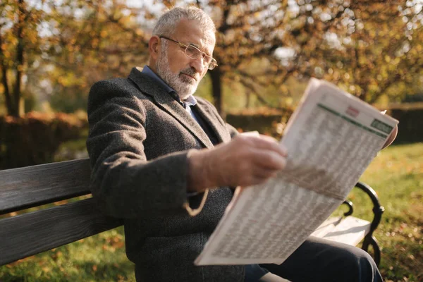 Jóképű nagyapa, gyönyörű szakállal, szürke kabátban ül egy padon a parkban, és újságot olvas. Idősebb ősz hajú férfi szemüvegben — Stock Fotó
