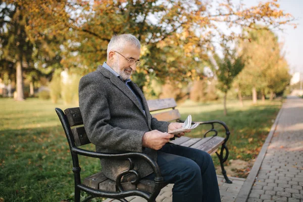 Skäggig äldre man i glasögon läser tidningen i höstparken. Snygg gråhårig man sitter på bänken tidigt på morgonen — Stockfoto
