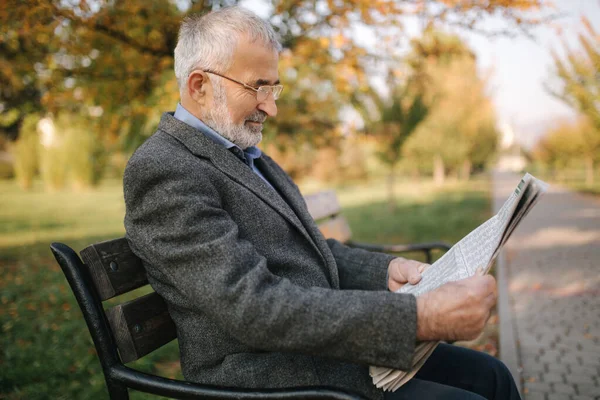 Hombre mayor barbudo en gafas leyendo periódico en el parque de otoño. Hombre guapo de pelo gris sentado en el banco temprano en la mañana — Foto de Stock