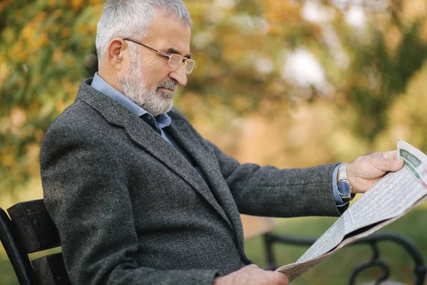 Homme âgé barbu dans des lunettes de lecture journal dans le parc d'automne. Beau homme aux cheveux gris assis sur le banc tôt le matin — Photo