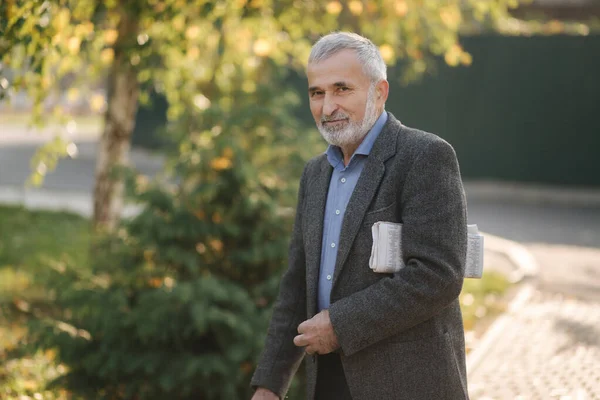 Un bell'uomo anziano passeggia per il parco con il giornale in mano. Barbuto nonno la mattina presto a piedi nel parco — Foto Stock