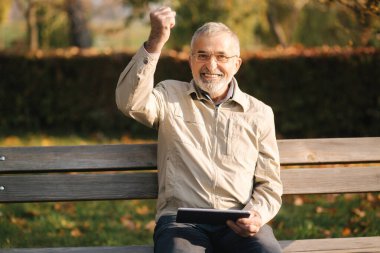 Bankta oturan yakışıklı yaşlı adam internette kaydırma yapmak için tablet kullanıyor. Sonbahar ağacının arkaplanı