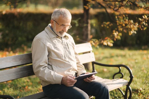 Hombre mayor guapo sentado en el banco y utilizar la tableta para desplazarse en Internet. Fondo del árbol de otoño —  Fotos de Stock