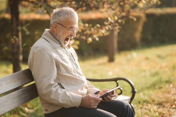 Όμορφος ηλικιωμένος που κάθεται στον πάγκο και να χρησιμοποιήσετε δισκίο για κύλιση στο διαδίκτυο. Ιστορικό φθινοπώρου δέντρου — Φωτογραφία Αρχείου