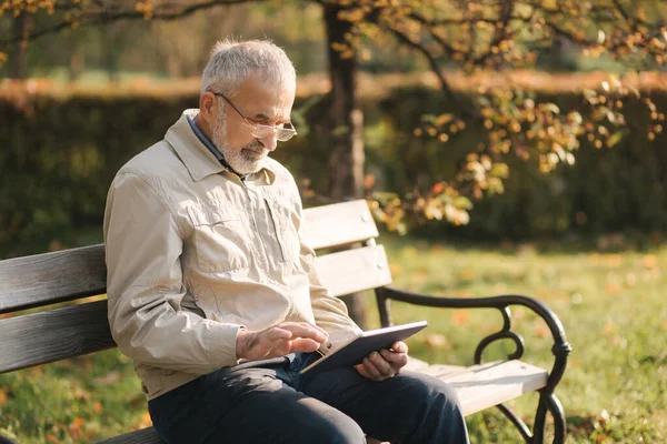 Hombre mayor guapo sentado en el banco y utilizar la tableta para desplazarse en Internet. Fondo del árbol de otoño —  Fotos de Stock