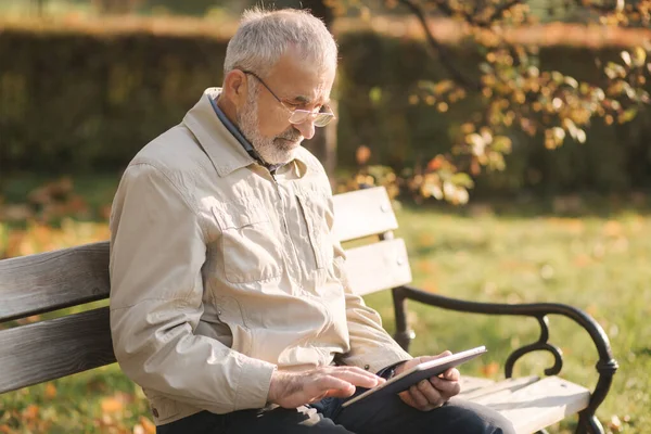 Beau vieil homme assis sur le banc et utiliser une tablette pour faire défiler Internet. Contexte de l'arbre d'automne — Photo