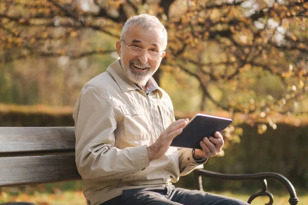 英俊的老人坐在长椅上，用平板电脑在网上滚动。 秋树的背景 — 图库照片