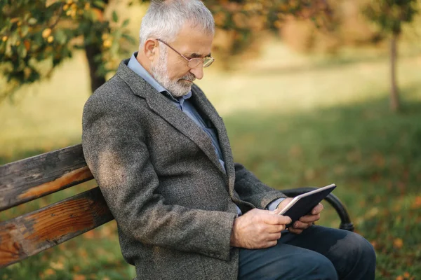 Красивый седой пожилой мужчина с красивой бородой сидит на скамейке и использовать планшет для прокрутки в Интернете. Фон осеннего дерева — стоковое фото