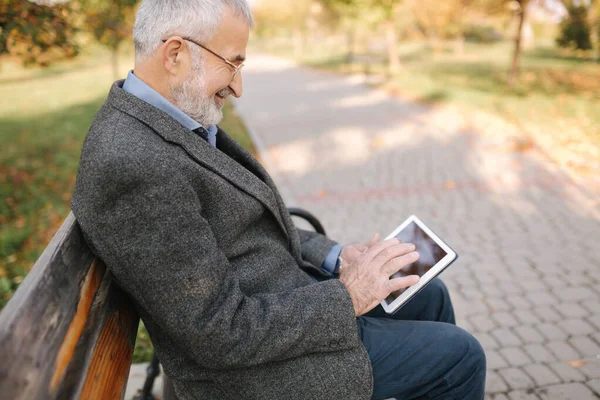 Handsome gray-haired elderly man with beautiful beard sitting on the bench and use tablet for scrolling in internet. Background of autumn tree