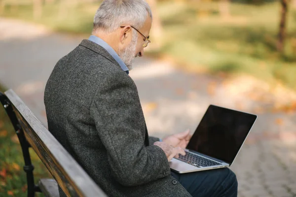 老人在公园里使用笔记本电脑。一个戴眼镜的英俊老人在外面工作 — 图库照片