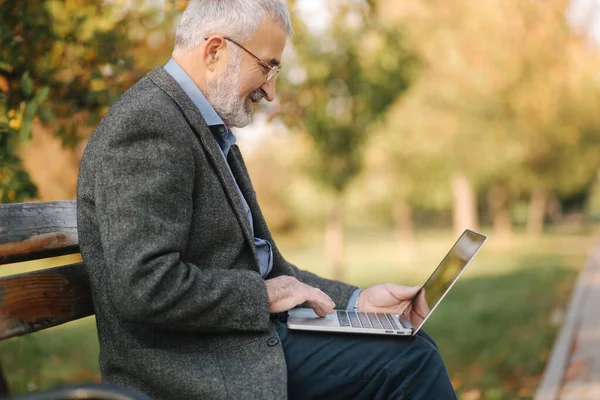 Äldre man använder laptop i parken. Stilig senior man i glasögon arbete utanför — Stockfoto