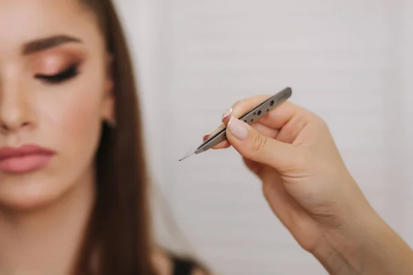 Close up view of make-up artist puts false eyelashes using tweezers. Background of white folding screen — Stock Photo, Image