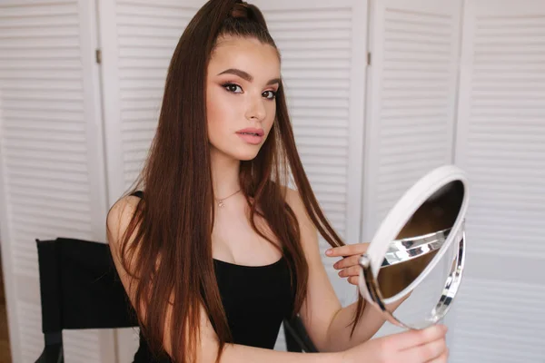 Young beautiful model looking in the mirror after makeup. Attractive young woman sitting in the beauty studio. Background of white folding screen — Stock Photo, Image