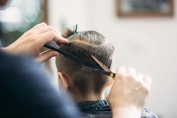 Pequeño chico consiguiendo corte de pelo por peluquero mientras está sentado en silla en la barbería . — Foto de Stock