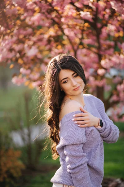 Attraktive junge Frau im lila Pullover steht vor einem Sakura-Baum im Park — Stockfoto