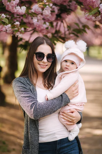 O mamă frumoasă cu fetiţa ei. Mama în ochelari de soare. Fundalul copacului roz — Fotografie, imagine de stoc