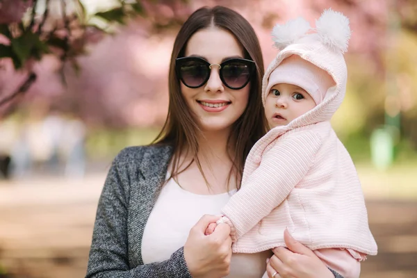 Beautiful mom with her little baby girl. Mom in sunglasses. Background of pink tree — Stock Photo, Image
