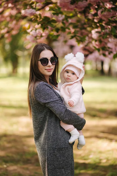 O mamă frumoasă cu fetiţa ei. Mama în ochelari de soare. Fundalul copacului roz — Fotografie, imagine de stoc