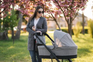 Parkta bebek arabası ile anne standı portresi. Mutlu genç anne bebekle yürüyor. Pembe ağacın arka planı