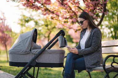 Bebek arabasında bebeğiyle bankta oturan ve telefonu kullanan genç bir anne. Mothe internetten bir şeye bak.