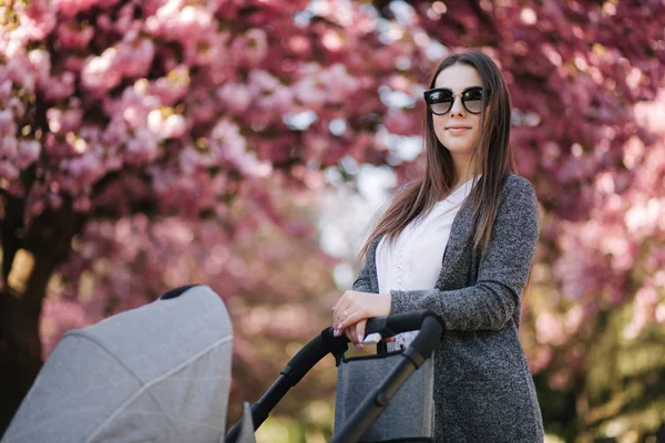 Retrato de mamá de pie con cochecito en el parque. Feliz madre joven caminando con el bebé. Fondo del árbol rosa —  Fotos de Stock