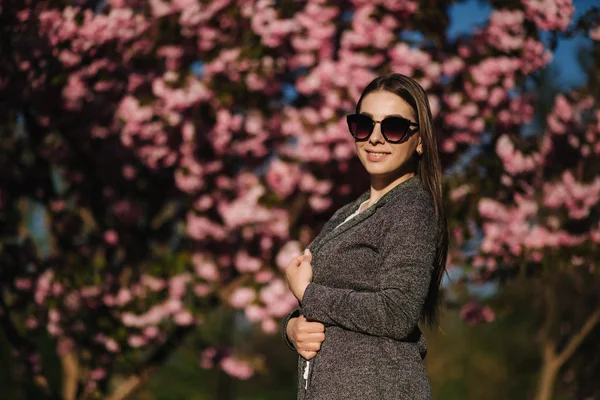 Hermosa chica en chaqueta y gafas de sol de pie junto al árbol de sakura. Hermoso árbol rosa en el parque —  Fotos de Stock