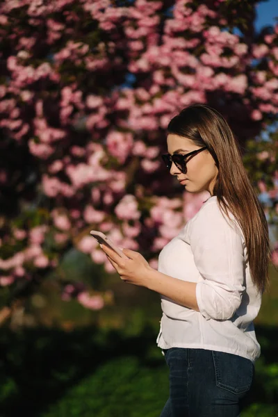 Jonge vrouw in wit shirt gebruik smartphone buiten. Achtergrond van de roze boom — Stockfoto