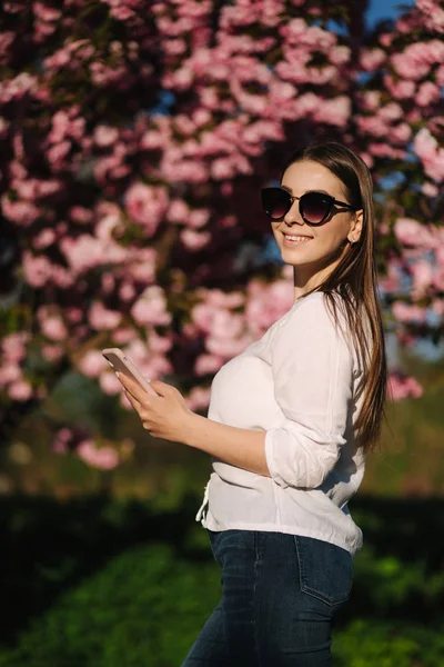 Mujer joven con camisa blanca usar teléfono inteligente fuera. Fondo del árbol rosa — Foto de Stock