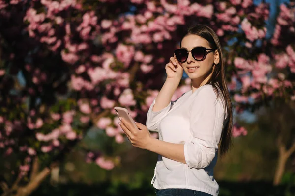 A fehér inges fiatal nő okostelefont használ odakint. A rózsaszín fa háttere — Stock Fotó