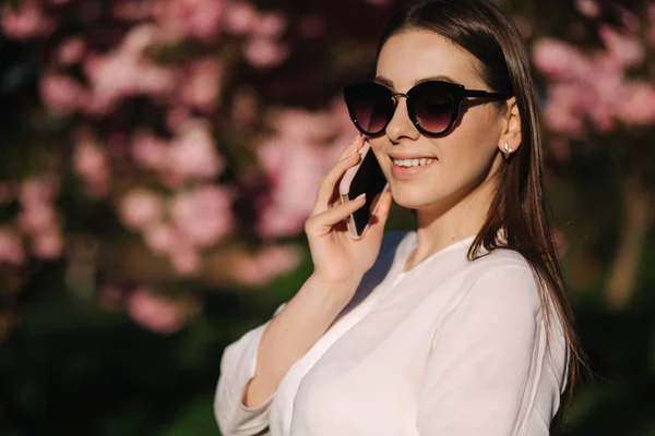 Retrato de mujer joven attravtive en camisa blanca y gafas de sol utilizan teléfono inteligente en el exterior. Habla con someboby por teléfono. Fondo del árbol rosa — Foto de Stock