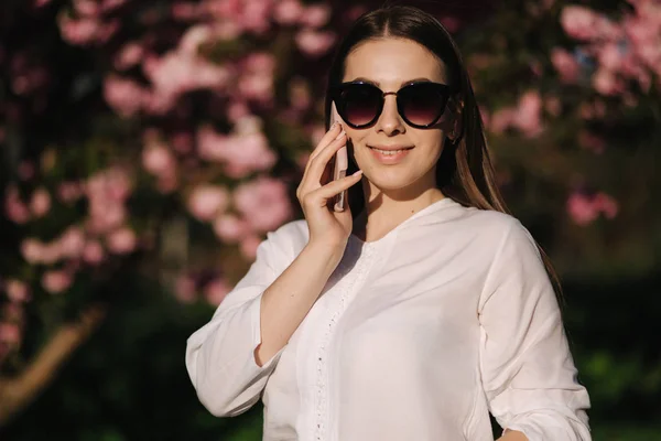 Retrato de mulher jovem attravtive em camisa branca e óculos de sol usar smartphone fora. Fala com alguém ao pé do telefone. Fundo da árvore rosa — Fotografia de Stock