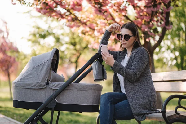 若いお母さんはベビーカーで赤ちゃんとベンチに座って電話を使います。インターネットで何かを見て — ストック写真