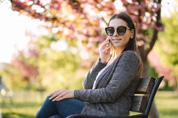 Hermosa mujer sentada en el banco y usando el teléfono. Mujer con estilo en el parque — Foto de Stock