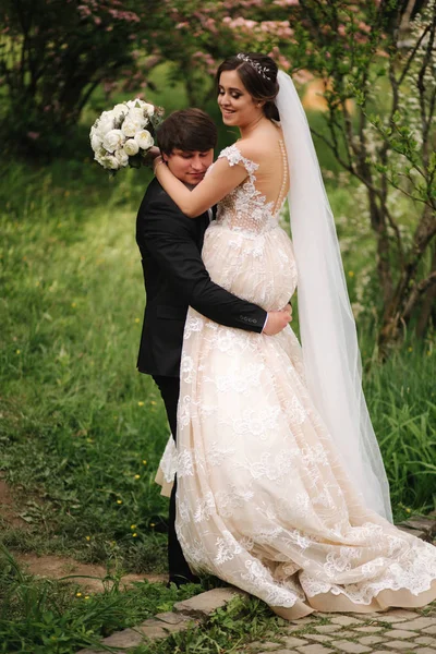 Stilig brudgummen kram hans vackra brud. Nygifta promenader i parken. Brun hår kvinna i elegant bröllopsklänning. Blossom Tree — Stockfoto