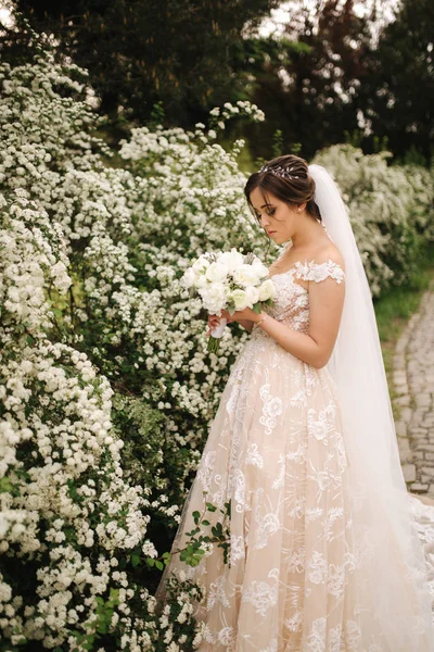 豪華なウェディングドレスで魅力的な花嫁は花束を保持し、咲く木の前に立ちます — ストック写真