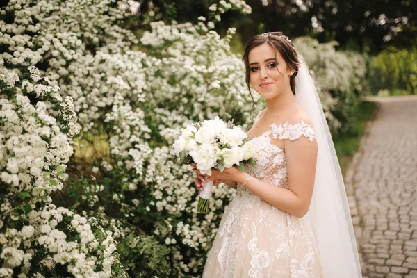 Noiva encantadora em lindo vestido de noiva segurar buquê e ficar na frente de árvores florescendo — Fotografia de Stock