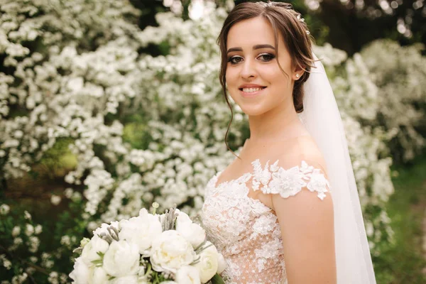Charmante Braut im wunderschönen Brautkleid hält Strauß und steht vor blühenden Bäumen — Stockfoto