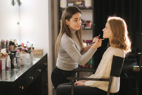 Beautiful female makeup artist doing makeup for a young blond hair girl in a beauty salon sitting in front of a large mirror. Concept of preparation for the holiday and meeting. Start working — Stock Photo, Image