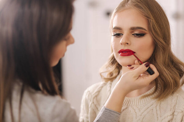Makeup artist applies red lipstick. Hand of make-up master, painting lips of young beauty blond hair model. Make up in process. Closeup view