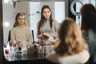 Güzel bir bayan makyöz, büyük bir aynanın önünde oturan sarışın bir kız için makyaj yapıyor. Tatil ve toplantı için hazırlık kavramı. Çalışmaya başla.