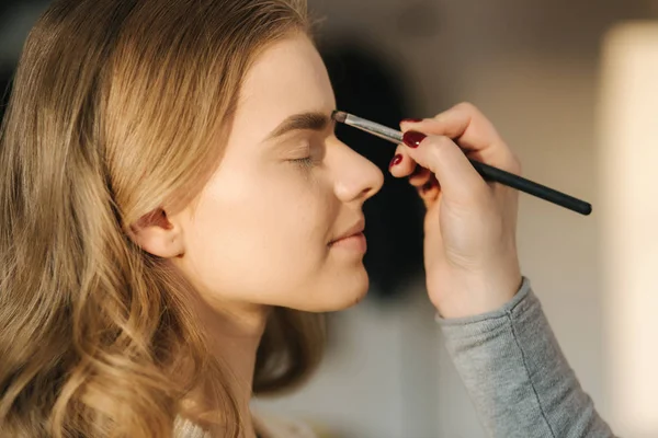 Makeup artist work in her beauty studio. Portrait of Woman applying by professional make up master. Beautiful make up artist start making a makeup for blond hair model
