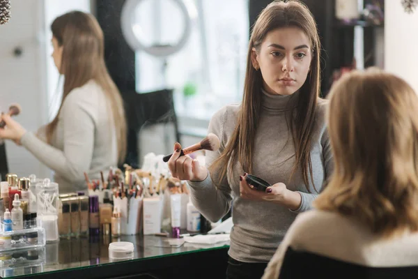 漂亮的女性化妆师在一个大镜子前的美容院为一个年轻的金发女孩化妆。准备假日和会议的概念.开始工作 — 图库照片