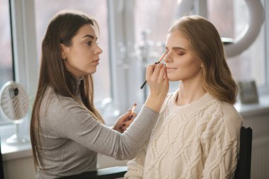 Makyaj sanatçısı güzellik salonunda çalışıyor. Profesyonel makyaj ustası tarafından uygulanan kadın portresi. Güzel makyözler sarı saç modeli için makyaj yapmaya başlar.