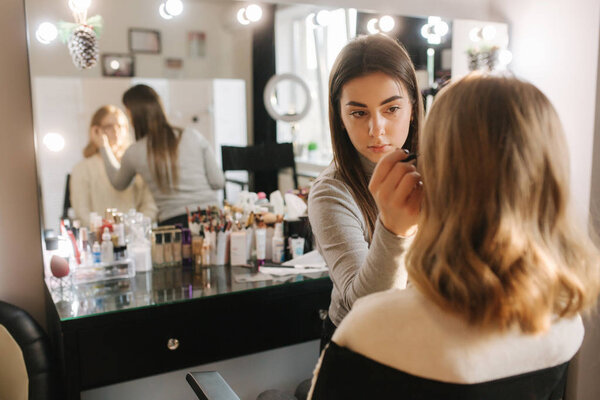 Beautiful female makeup artist doing makeup for a young blond hair girl in a beauty salon sitting in front of a large mirror. Concept of preparation for the holiday and meeting. Start working