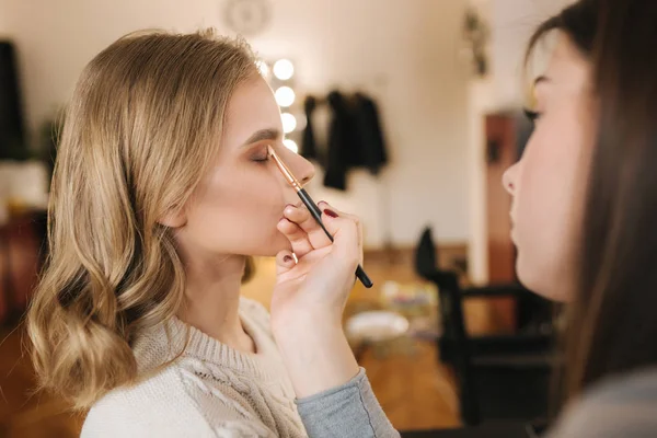 Makeup artist work in her beauty studio. Portrait of Woman applying by professional make up master. Beautiful make up artist start making a makeup for blond hair model