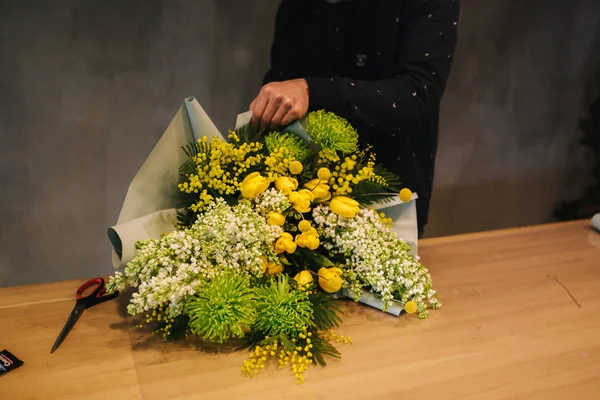 Macho hacer ramo de primavera. Hombre florista envolviendo hermoso ramo de flores de primavera en papel de paquete en la mesa de madera. Hermosa composición de flores amarillas —  Fotos de Stock