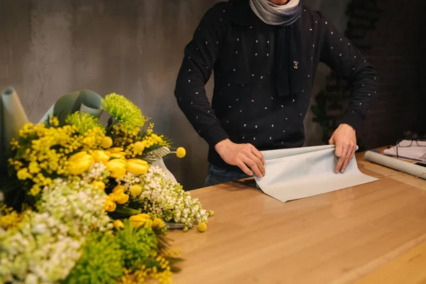 Een man maakt lenteboeket. Man bloemist wikkelen mooi boeket van de lente bloemen in pakpapier op de houten tafel. Prachtige bloemsamenstelling van gele bloemen — Stockfoto